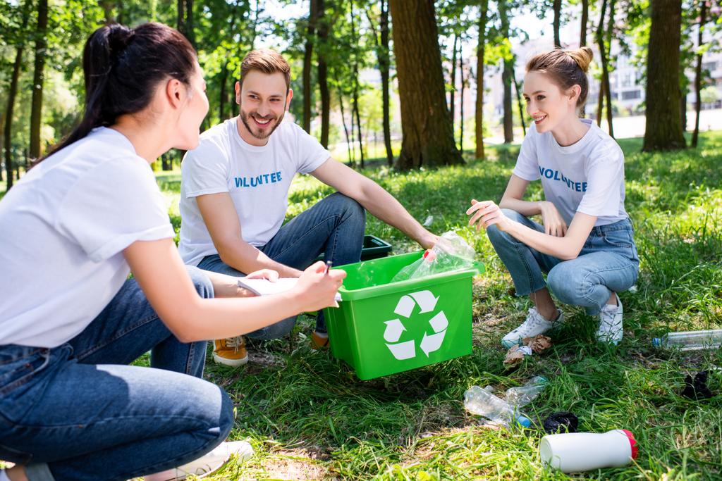 nuoret hymyilevät vapaaehtoiset, joilla on vihreä kierrätyslaatikko puistossa
 - Valokuva, kuva