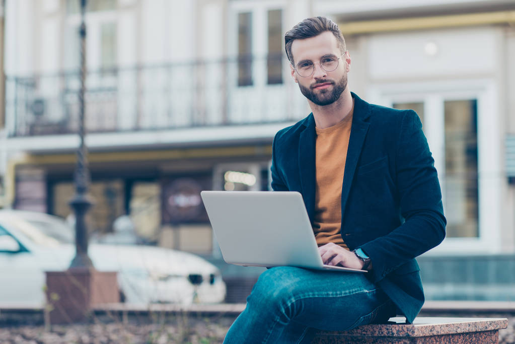 Портрет привлекательного элегантного джентльмена в джинсовой куртке, сидящего снаружи с помощью компьютера, смотрящего в камеру и наслаждающегося городской атмосферой
 - Фото, изображение