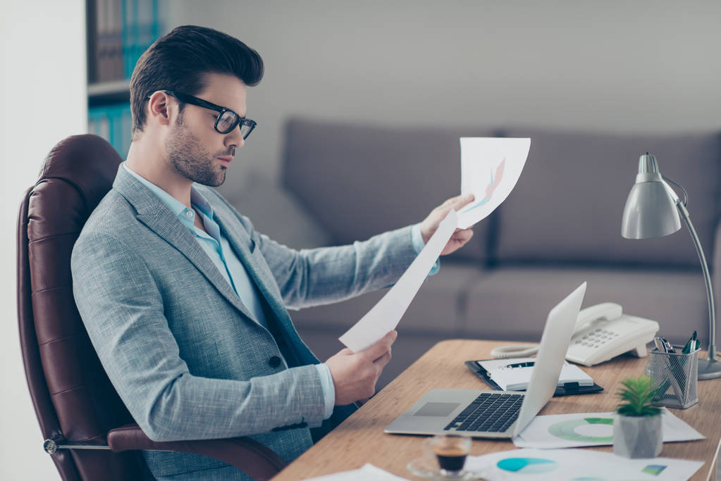 Portrait of thoughtful stylish financier holding papers in hand, expertising documents, working on start up, preparing presentation, sitting at desktop in work station - Photo, Image