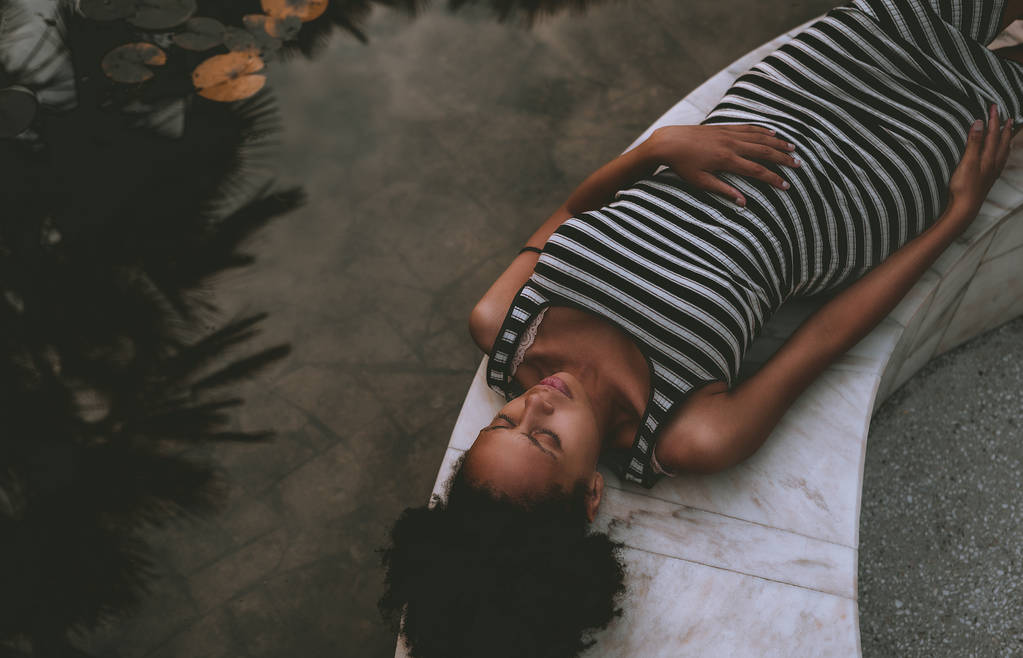 Joven mujer embarazada afroamericana en un vestido a rayas y con el pelo rizado Afro está descansando en el atardecer de verano en el banco de mármol al lado del estanque con nenúfares y reflejos de las palmeras
 - Foto, Imagen