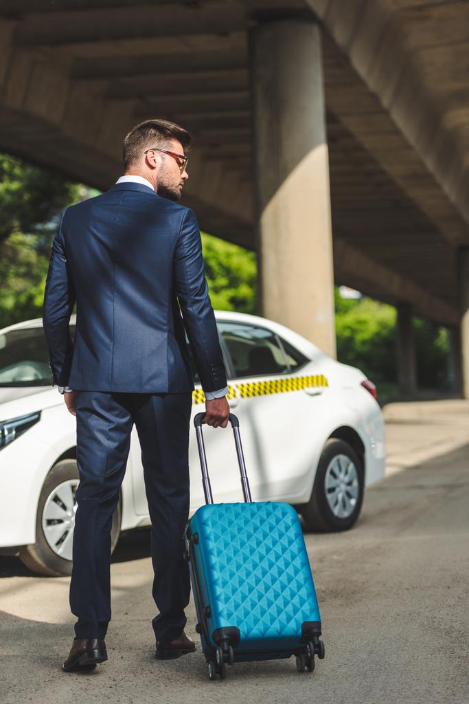 Achteraanzicht van stijlvolle jonge zakenman met koffer staande in de buurt van taxi - Foto, afbeelding