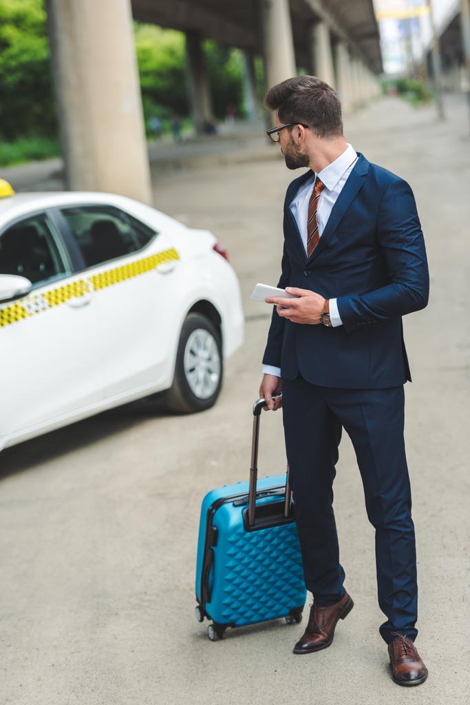 hombre guapo con maleta y teléfono inteligente mirando taxi en la calle
 - Foto, Imagen