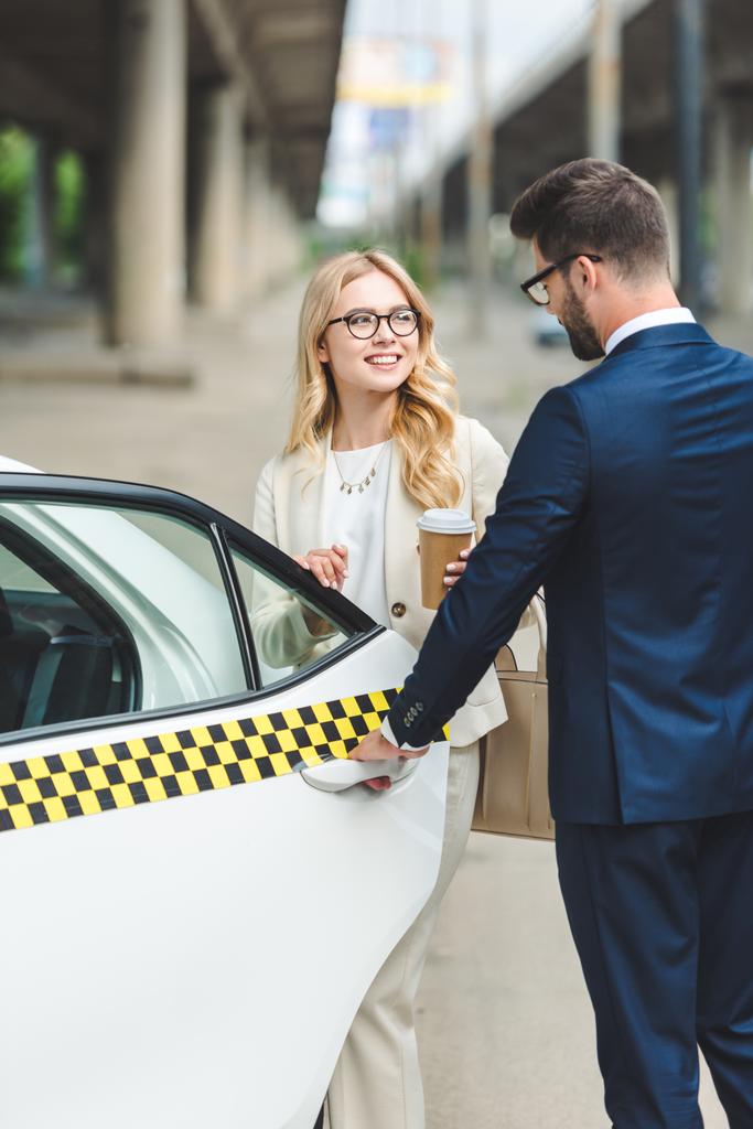 mooie glimlachend blond meisje bedrijf koffie te gaan en kijken naar knappe man openen van de deur van de taxi - Foto, afbeelding