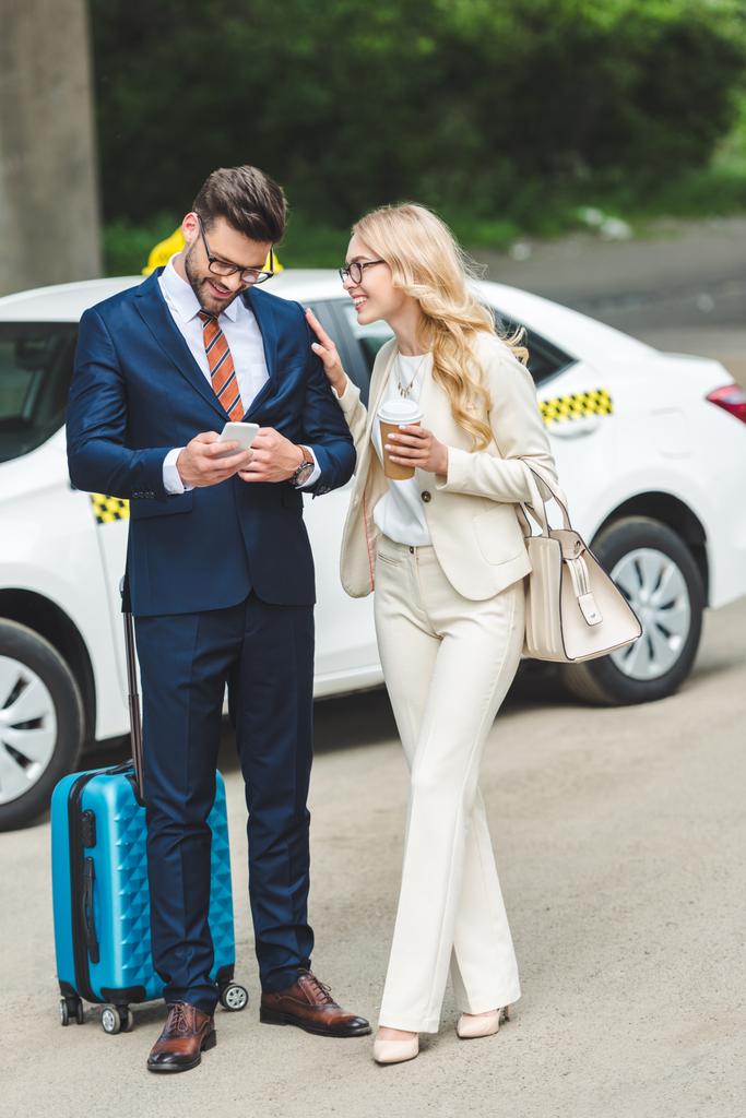 smiling blonde woman holding coffee to go and looking at handsome man using smartphone while standing with suitcase near taxi - Photo, Image