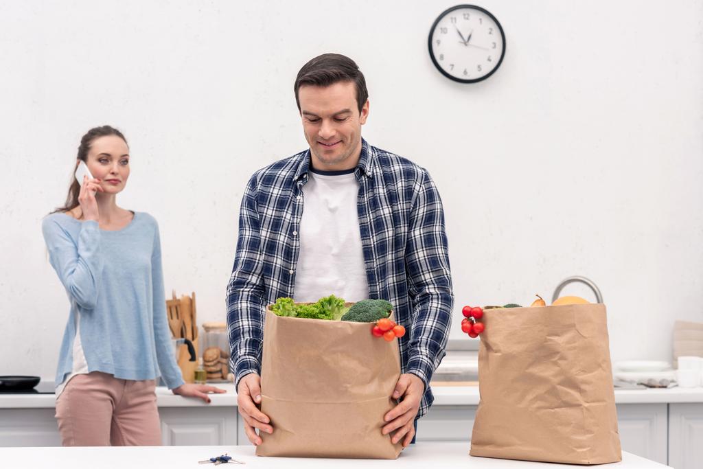 щасливий дорослий чоловік носить паперовий пакет з продуктового магазину, а його дружина розмовляє по телефону
 - Фото, зображення