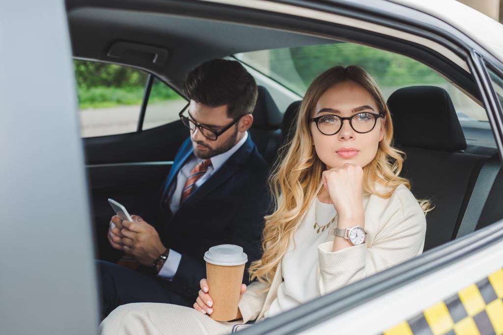 молода жінка тримає паперову чашку і дивиться геть, поки чоловік використовує смартфон у таксі
 - Фото, зображення