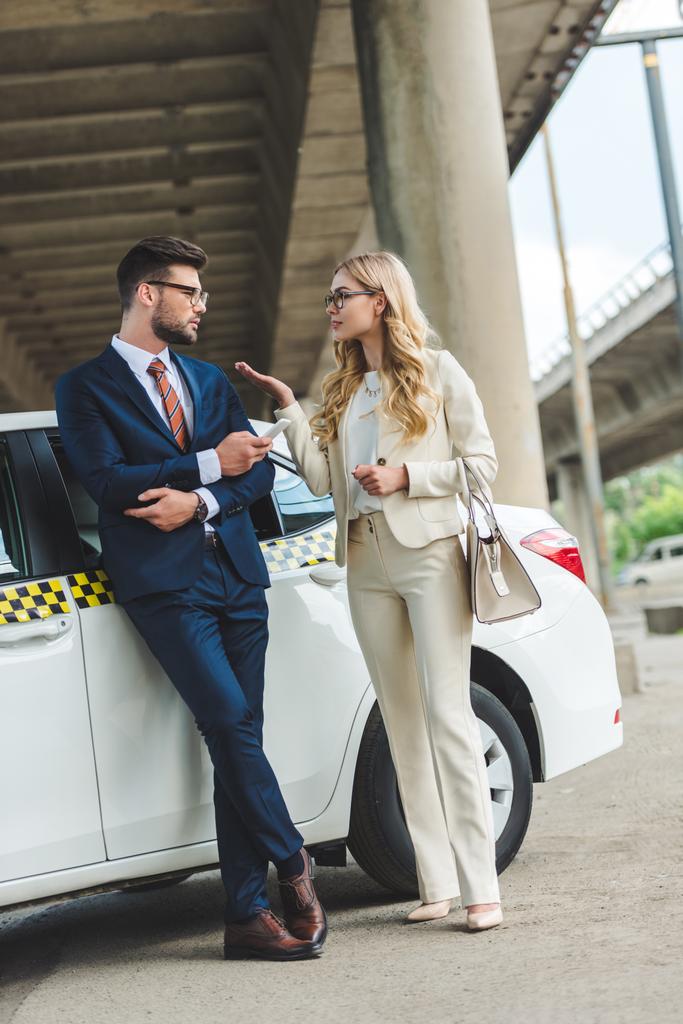 young couple in formal wear and eyeglasses talking and looking at each other while standing near taxi - Photo, Image