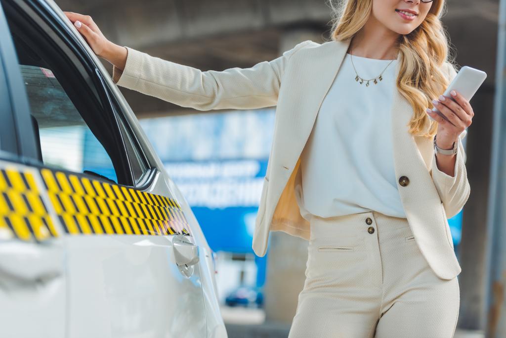 oříznuté záběr usměvavá blondýnka pomocí smartphone stoje u taxíku - Fotografie, Obrázek