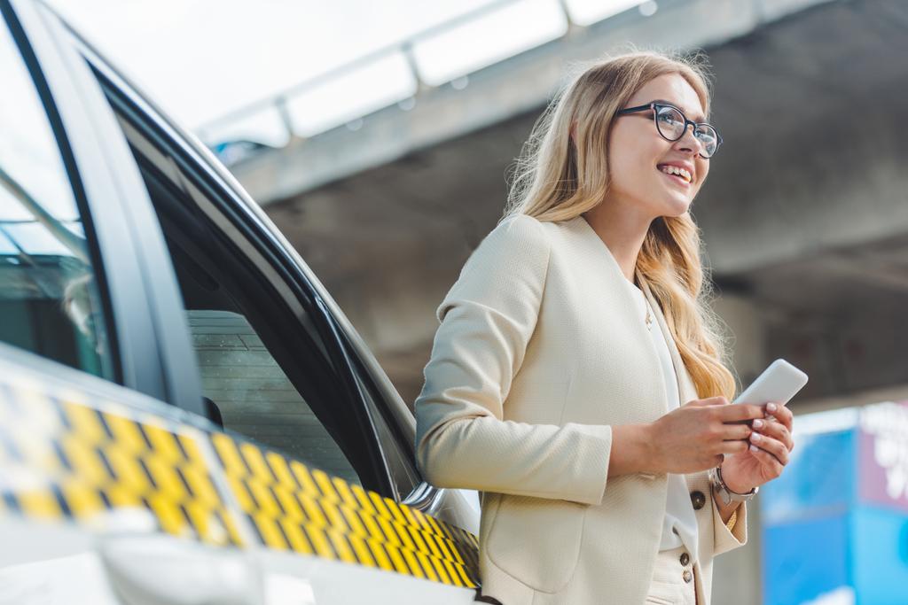 Tiefansicht eines lächelnden blonden Mädchens mit Brille, das sein Smartphone in der Hand hält und wegschaut, während es in der Nähe des Taxis steht - Foto, Bild