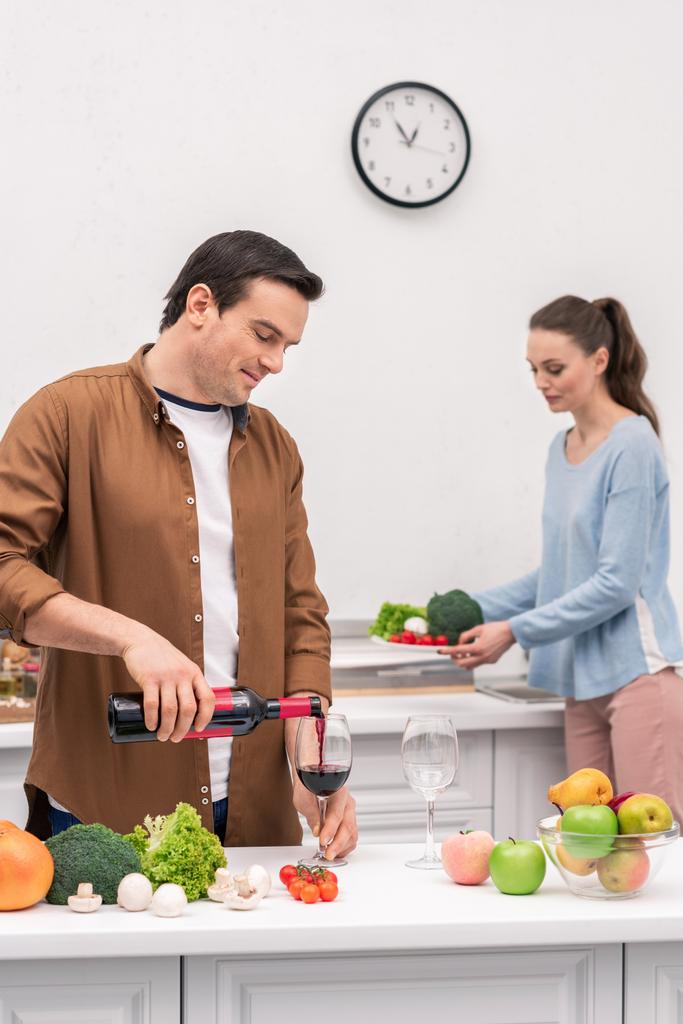 glücklicher Ehemann schenkt Frau während der Zubereitung des Abendessens Wein ein - Foto, Bild