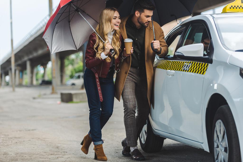 χαμογελαστός, νεαρό ζευγάρι με ομπρέλες βλέπουν αυτοκίνητο ταξί - Φωτογραφία, εικόνα