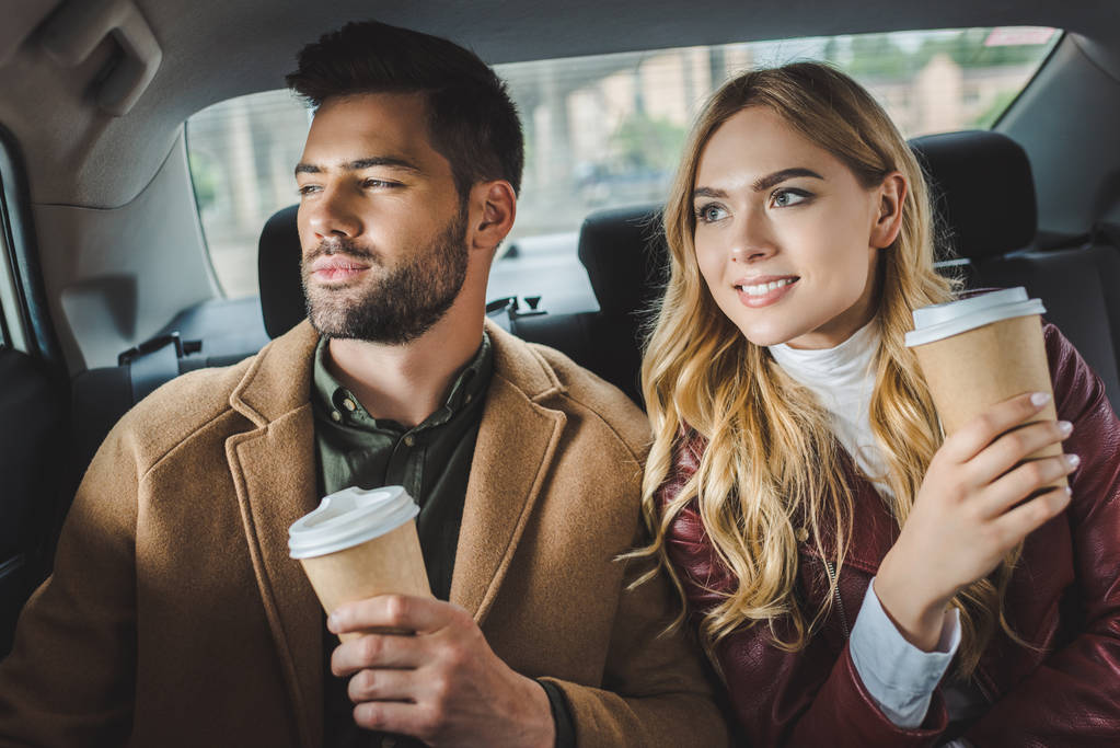 Улыбающаяся стильная молодая пара с бумажными стаканчиками, сидящая вместе в такси
 - Фото, изображение