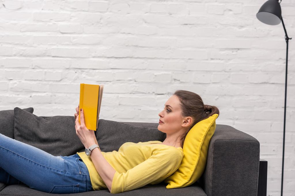 вид сбоку на взрослую женщину, читающую книгу, лежа на диване дома
 - Фото, изображение