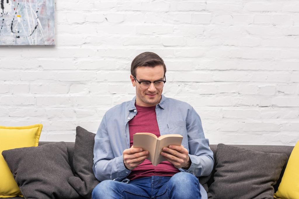 happy adult man reading book on sofa at home in front of white brick wall - Photo, Image