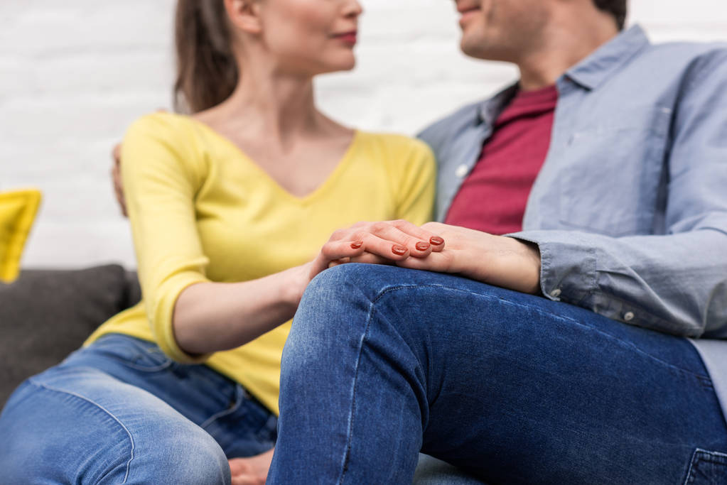 abgeschnittene Aufnahme eines erwachsenen Paares, das Händchen hält, während es zu Hause auf der Couch sitzt - Foto, Bild
