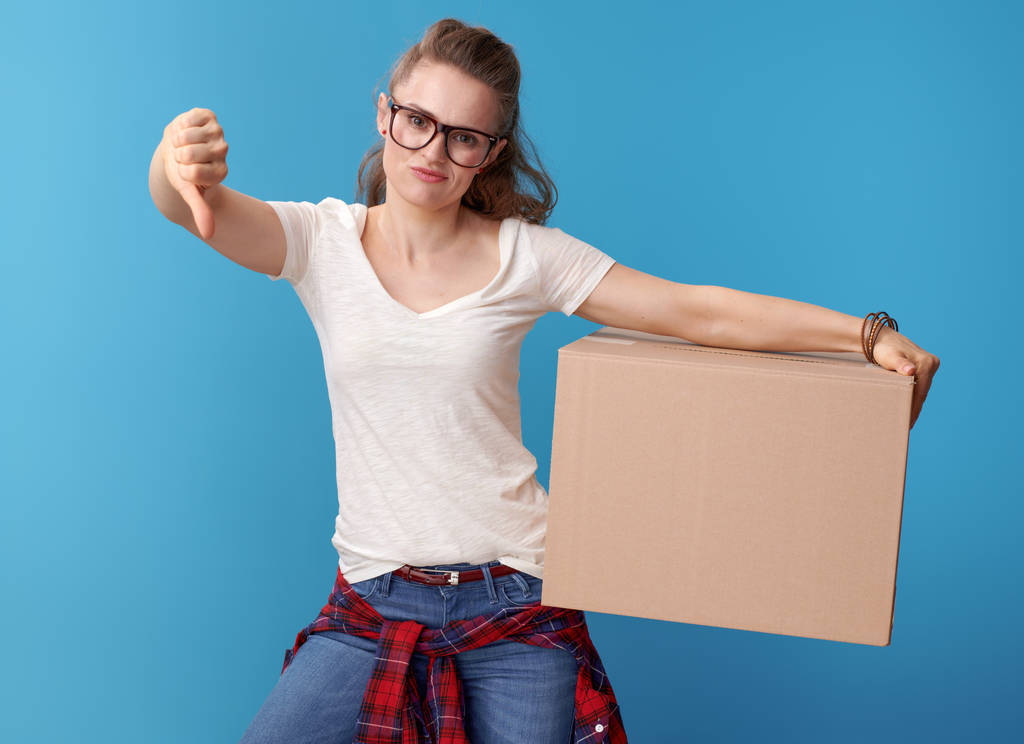 hipster moderno infeliz en camisa blanca con una caja de cartón que muestra los pulgares hacia abajo sobre fondo azul
 - Foto, imagen