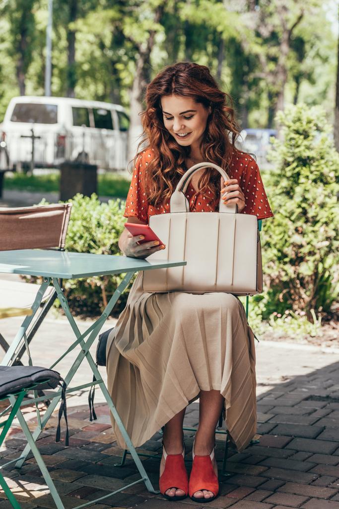 κομψή γυναίκα με τσάντα χρησιμοποιώντας smartphone, ενώ κάθεται στην βεράντα στο καφέ - Φωτογραφία, εικόνα