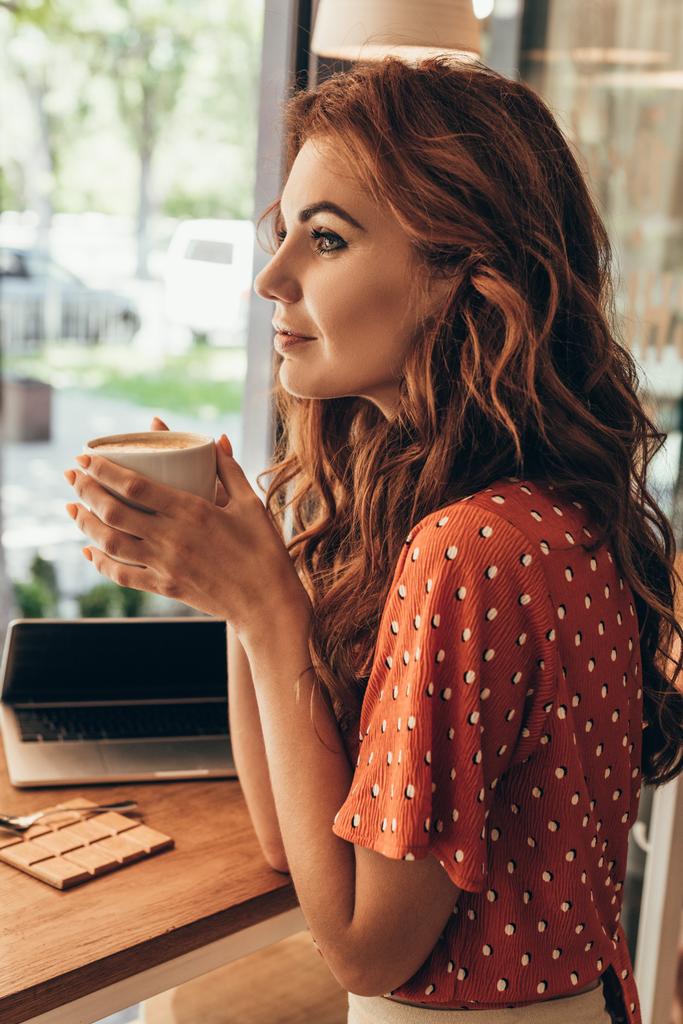 vista lateral da mulher pensativa com xícara de café à mesa com laptop no café
 - Foto, Imagem