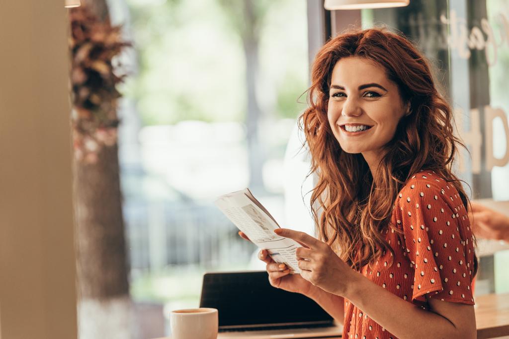oldalnézet, mosolygós nő újság asztalnál kezében laptop a kávézóban - Fotó, kép