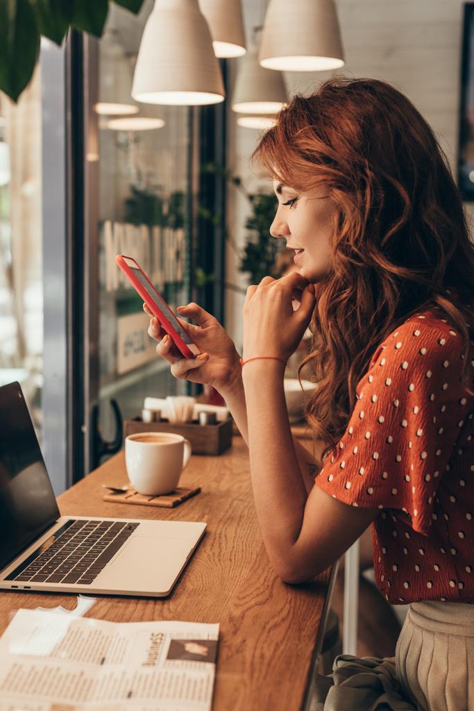 a fiatal nő használ smartphone asztalhoz a laptop a kávézóban oldalnézete - Fotó, kép