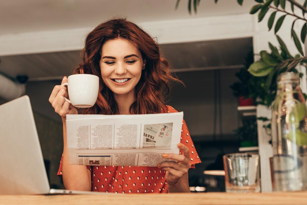 Πορτρέτο γυναίκας χαμογελαστά με φλιτζάνι καφέ διαβάζοντας εφημερίδα στο τραπέζι με το φορητό υπολογιστή στο κατάστημα καφέ - Φωτογραφία, εικόνα