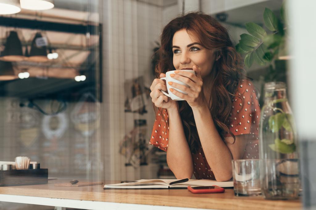 Portret van een jonge vrouw die koffie drinkt aan tafel met notebook in café - Foto, afbeelding