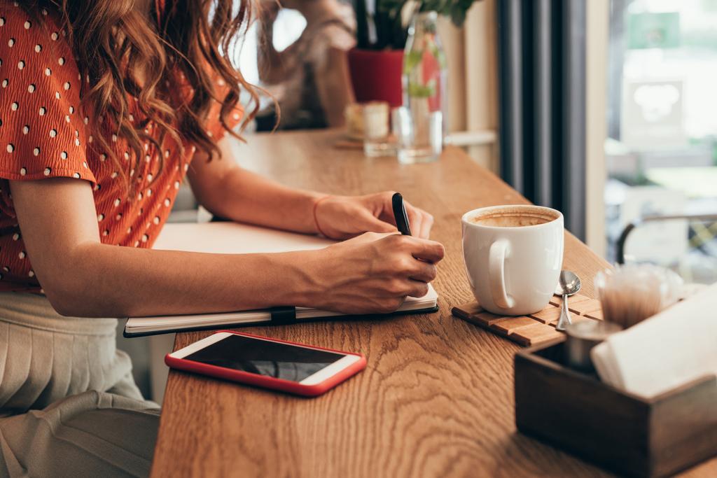 recortado tiro de bloguero haciendo notas en el cuaderno en la mesa con taza de café en la cafetería
 - Foto, imagen