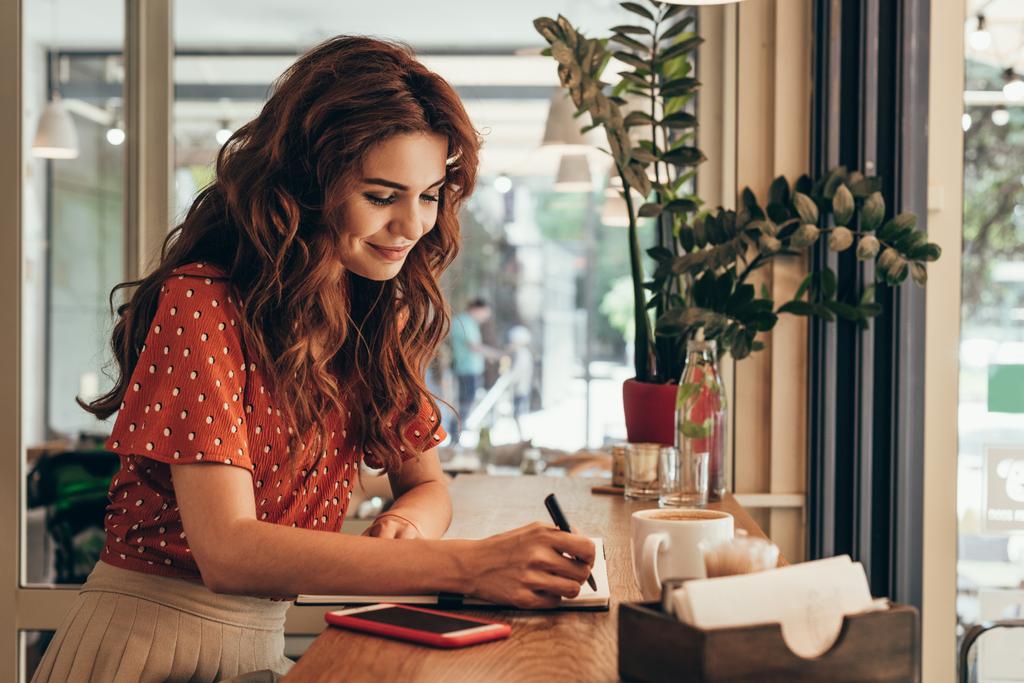 Seitenansicht einer jungen Bloggerin, die Notizen in Notizbuch am Tisch mit einer Tasse Kaffee im Café macht - Foto, Bild