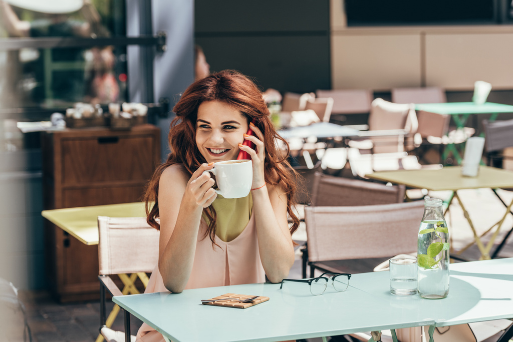 Portret van lachende vrouw met kopje koffie praten over smartphone in café - Foto, afbeelding