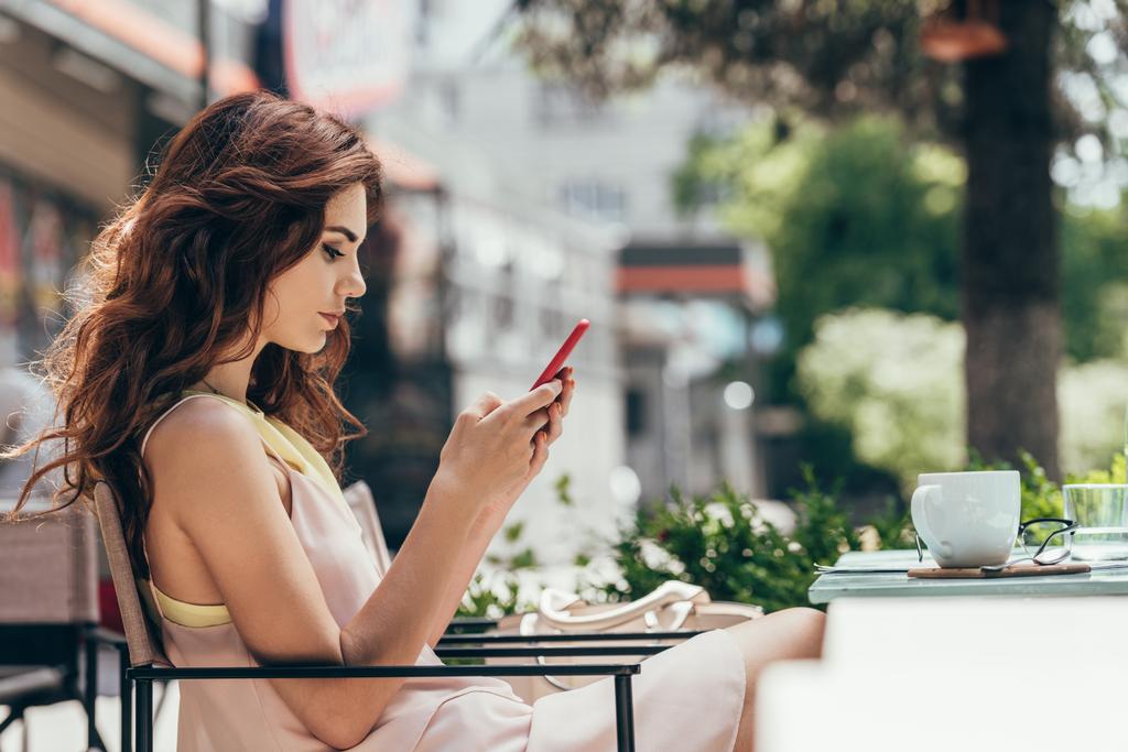 Seitenansicht einer jungen Frau, die im Café mit einer Tasse Kaffee am Tisch sitzt und ihr Smartphone benutzt - Foto, Bild