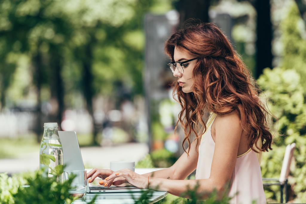 oldalnézetből a koncentrált fiatal blogger a szemüvegek dolgozik a laptop a kávézóban - Fotó, kép