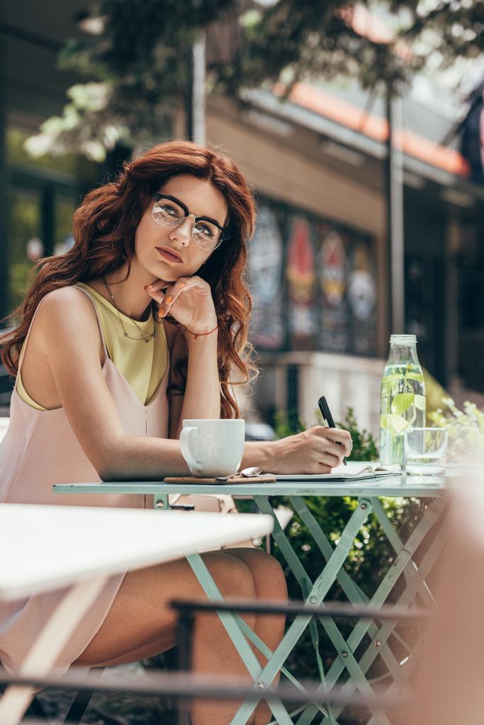 empresária pensativa em óculos sentados à mesa com caderno e xícara de café no restaurante
 - Foto, Imagem