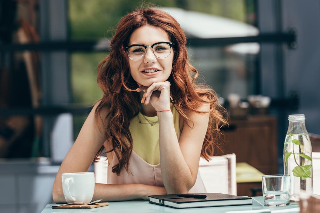 Portret van aantrekkelijke zakenvrouw in brillen zit aan tafel met kopje koffie en laptop in café - Foto, afbeelding