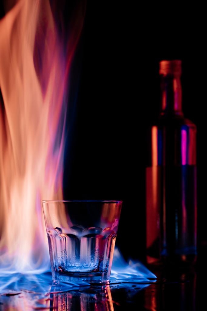 κλείνω πάνω θέα της φλόγας, κενό γυαλί και μπουκάλι sambuca αλκοολούχου ποτού σε μαύρο φόντο - Φωτογραφία, εικόνα