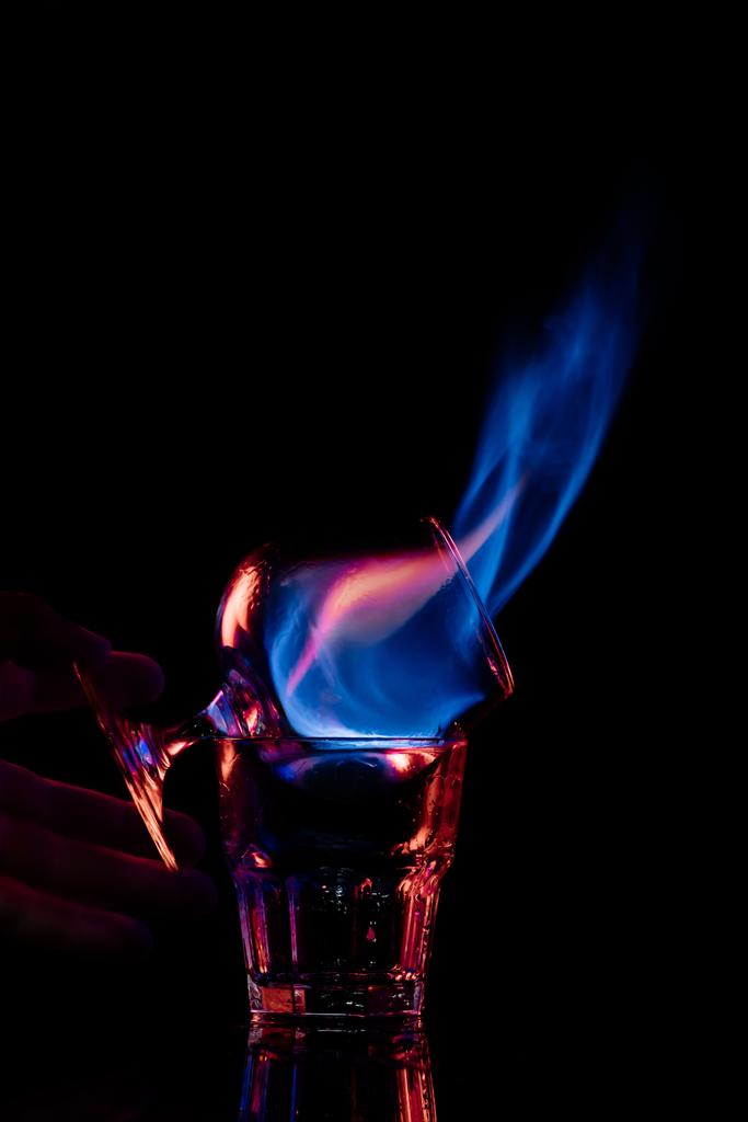 黒い背景にサンブカ アルコールを燃焼でガラスを保持している女性の部分的なビュー - 写真・画像