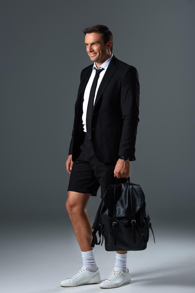 glimlachend stijlvolle man in korte broek holding rugzak op grijze achtergrond  - Foto, afbeelding