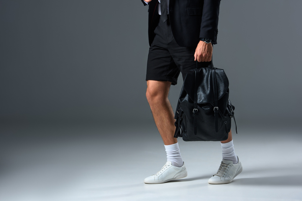 обрізаний образ стильного чоловіка в шортах, що тримає рюкзак на сірому фоні
  - Фото, зображення