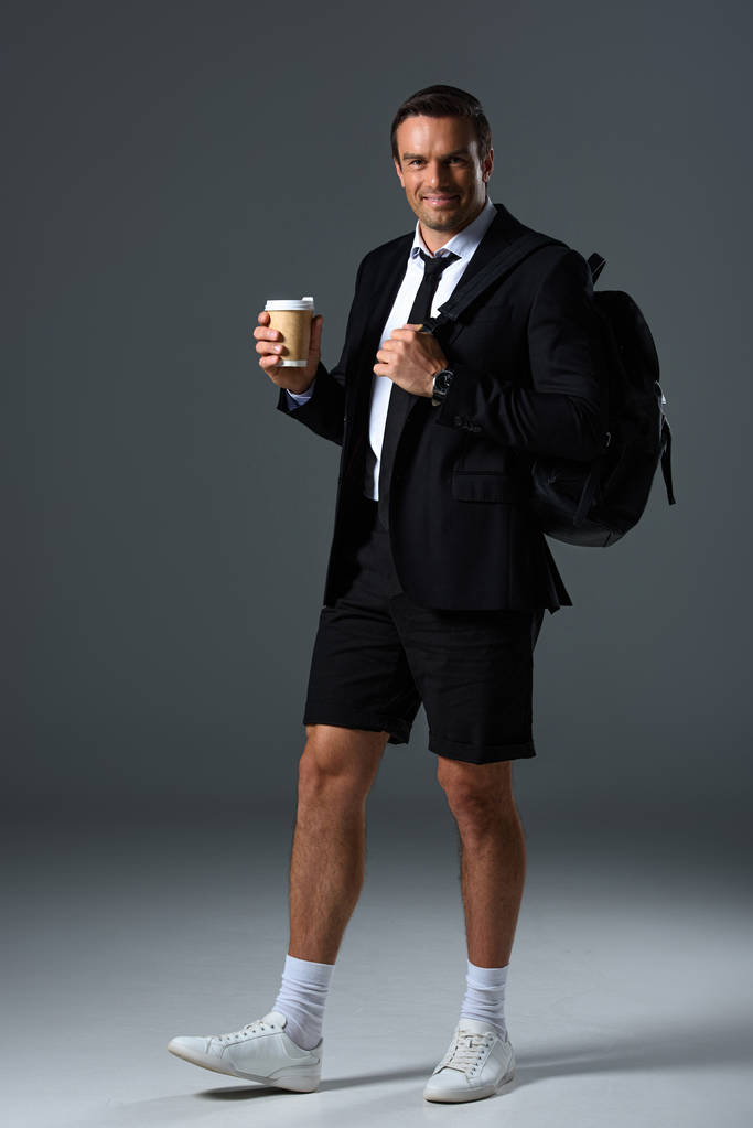 hombre con estilo feliz en pantalones cortos con mochila sosteniendo taza de papel de café sobre fondo gris
  - Foto, Imagen