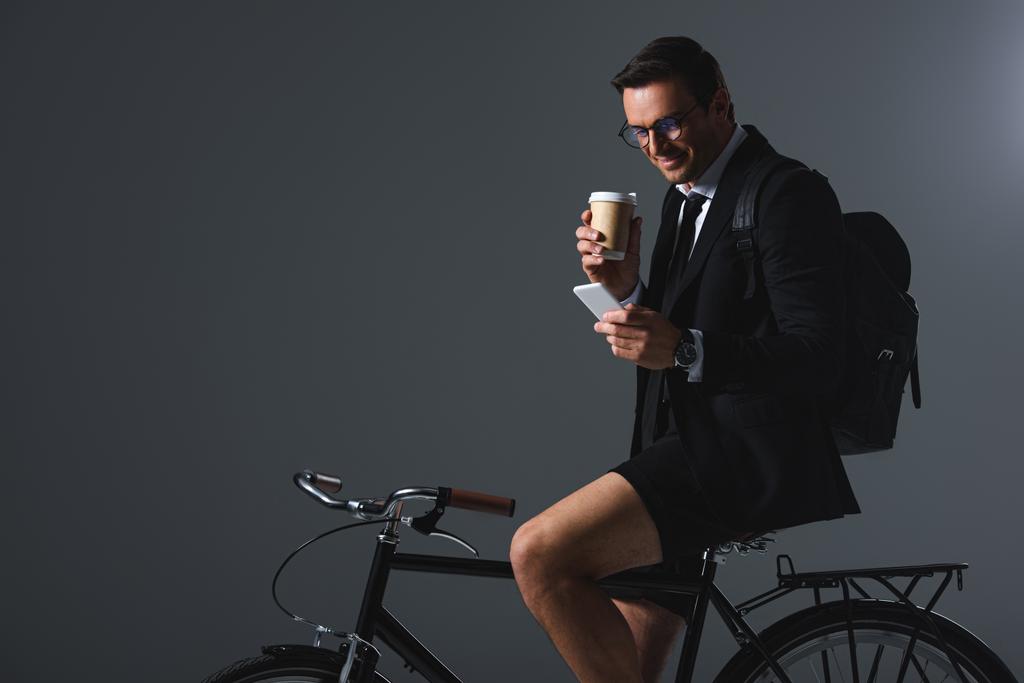 κομψό άνδρα με σακίδιο κάθεται στο ποδήλατο με έξυπνο κινητό τηλέφωνο στο χέρι και πίνοντας καφέ σε γκρι φόντο  - Φωτογραφία, εικόνα