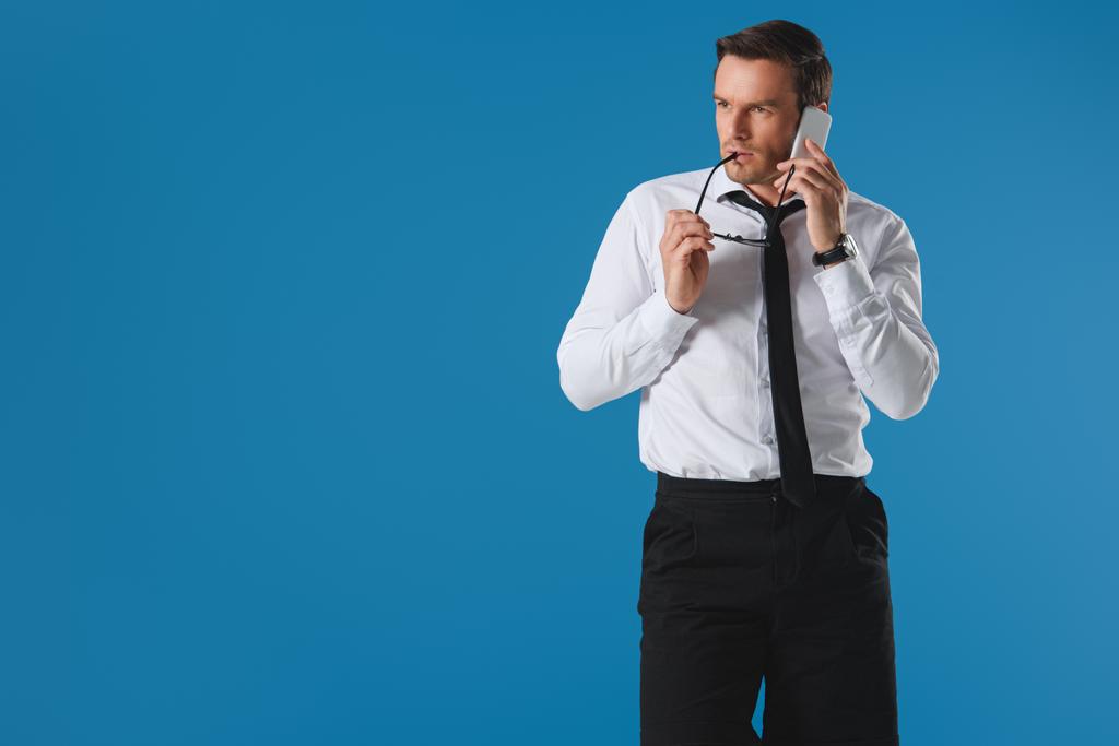 ernster Mann beißt Brille, während er mit dem Smartphone spricht und isoliert auf blauem Grund wegschaut - Foto, Bild
