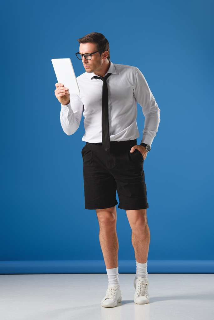 koncentruje się człowiek w okularach i szorty za pomocą cyfrowego tabletu stojąc z ręką w kieszeni na niebiesko - Zdjęcie, obraz