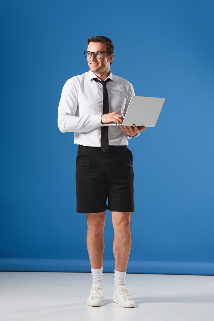 komea hymyilevä liikemies shortseissa kannettavan tietokoneen avulla ja katselee pois sinisellä
 - Valokuva, kuva