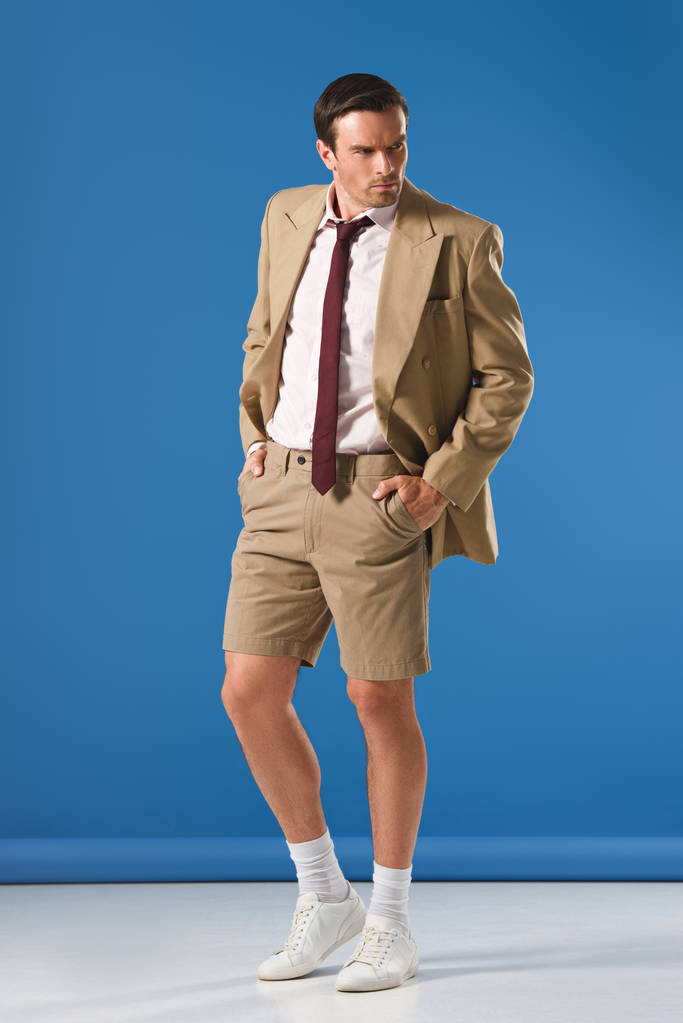 Ganzkörperansicht eines gutaussehenden Mannes in Anzughose und Shorts, der mit den Händen in den Taschen steht und wegschaut auf blau - Foto, Bild