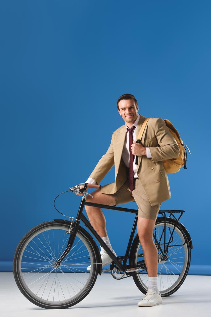 pohledný muž s batohem sedí na kole a usmívá se na kameru na modré - Fotografie, Obrázek