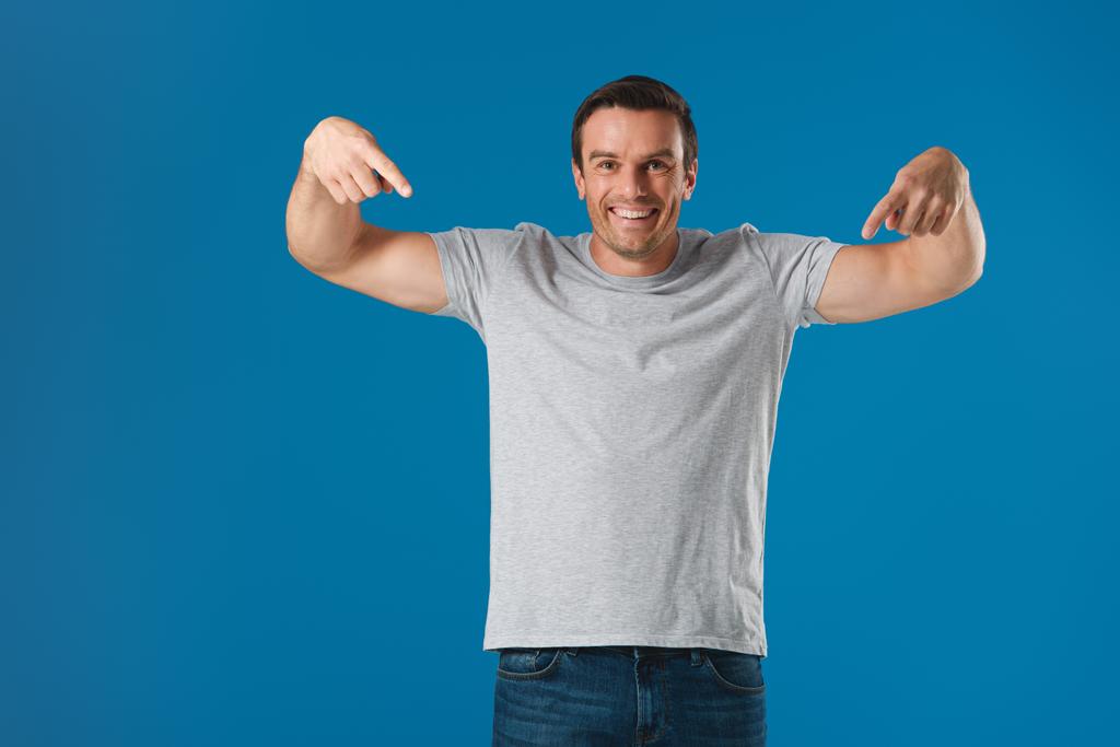 gelukkig man met vingers en t-shirt aan te wijzen en glimlachend op camera geïsoleerd op blauw - Foto, afbeelding