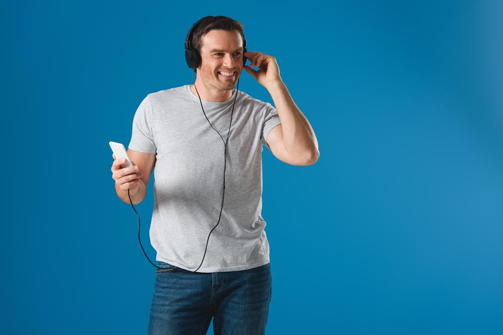 felice uomo in cuffia ascoltare musica con smartphone e distogliere lo sguardo isolato sul blu
 - Foto, immagini