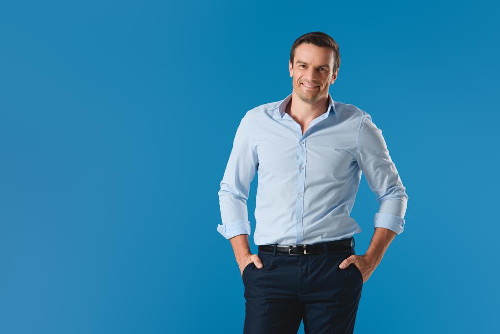 handsome man standing with hands in pockets and smiling at camera isolated on blue - Photo, Image