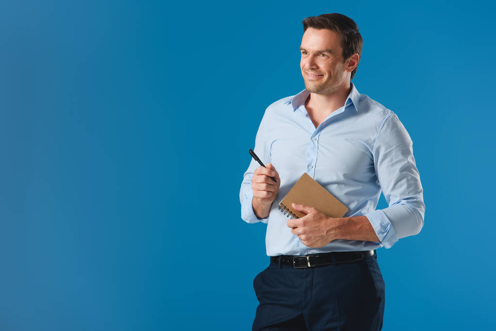 Красивый улыбающийся бизнесмен, держащий в руках блокнот и ручку, отводящий взгляд на синий
 - Фото, изображение