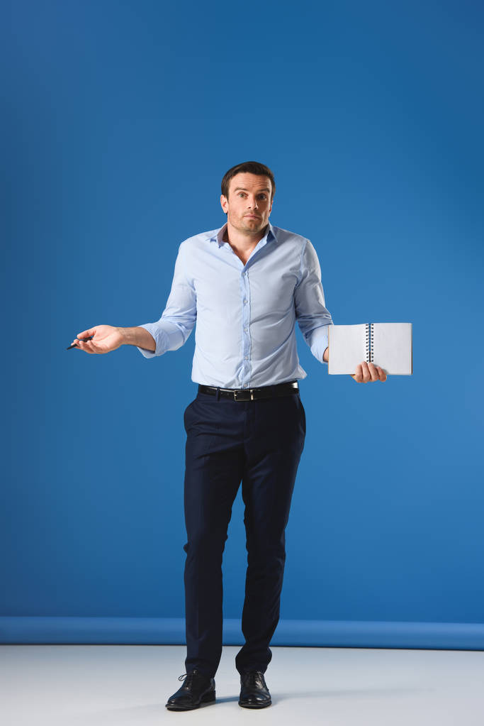full length view of man holding blank notebook and looking at camera on blue - Photo, Image