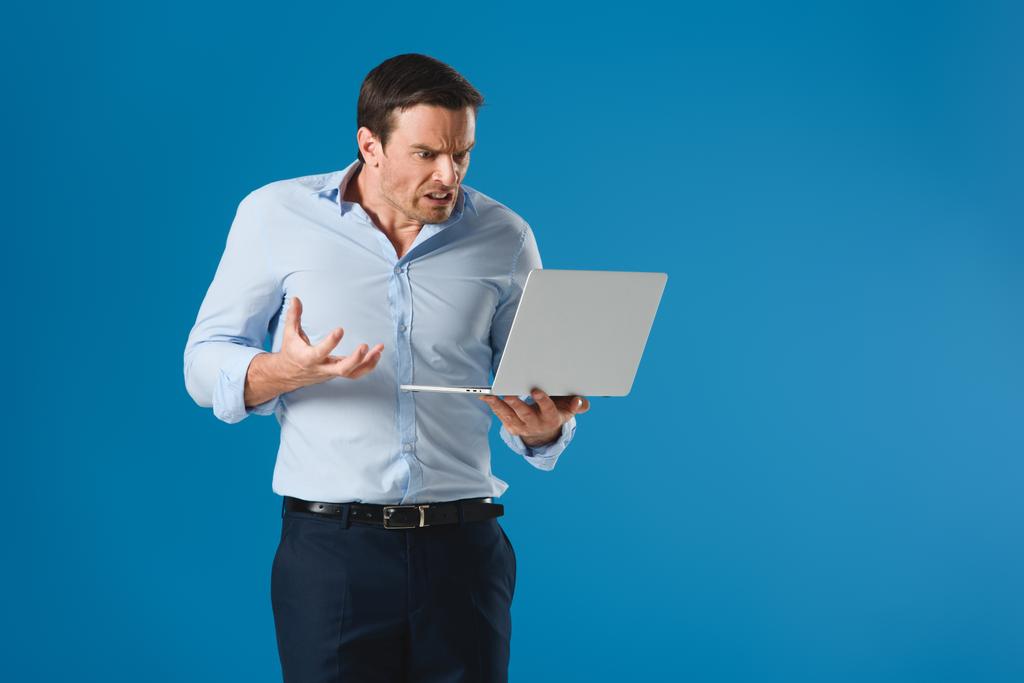 hombre enojado sosteniendo y usando el ordenador portátil aislado en azul
 - Foto, Imagen